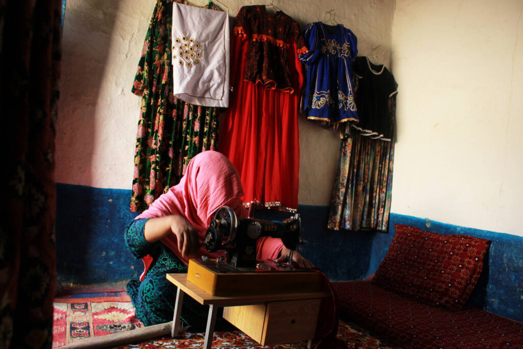 Gulmeena works at her sewing machine while the clothing she already has made hangs behind her at her home in the refugee village of Padhana, Pakistan.