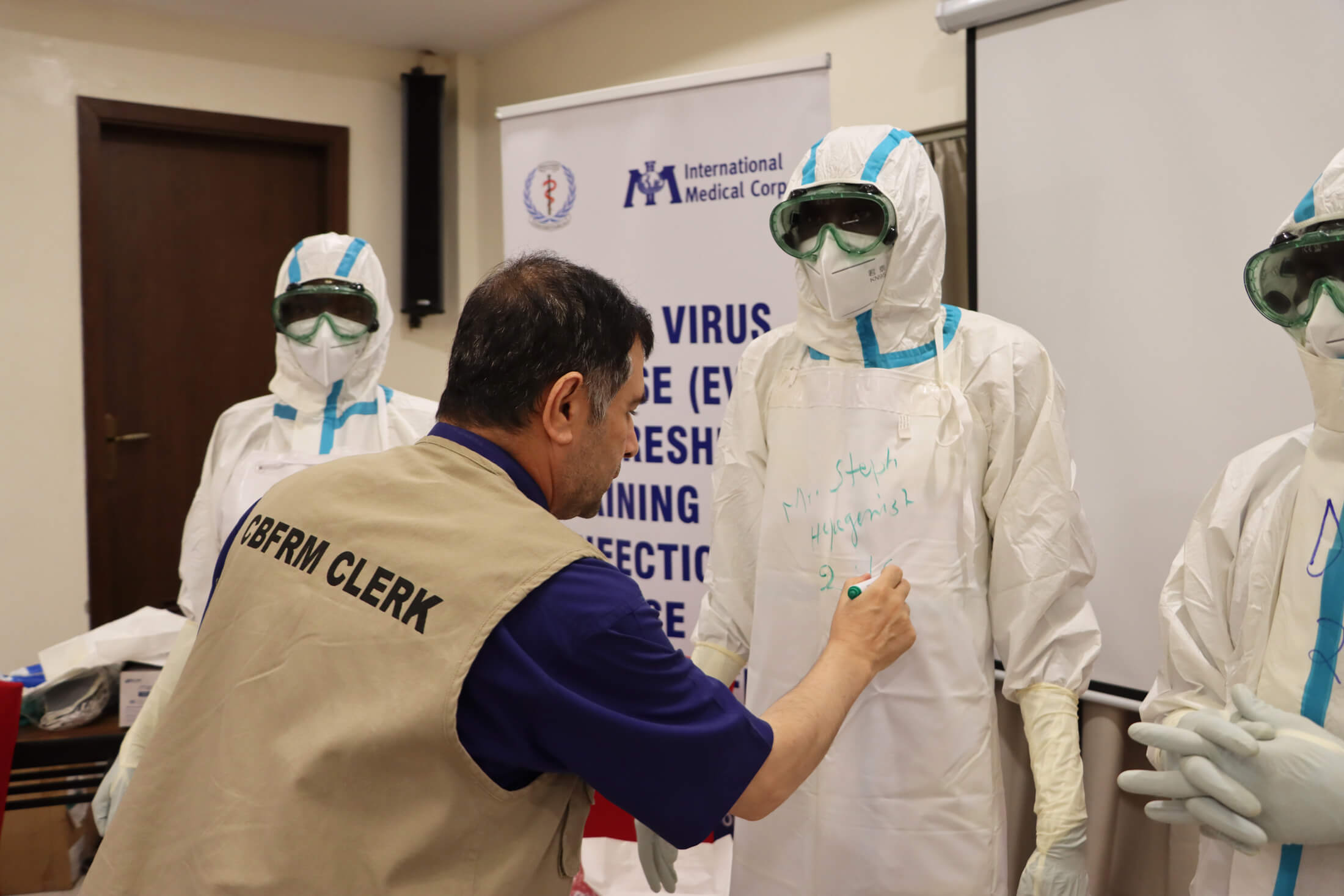 During a roleplaying exercise, Dr. Ebadullah Hedayat—who is playing a community‑based feedback and response mechanism clerk—writes the name of a health worker and the time the PPE was worn on the worker’s gown.